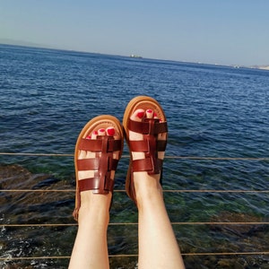 May include: A pair of brown leather sandals with a criss-cross strap design. The sandals are worn by a person with red nail polish on their toes. The person is standing on a wooden platform with a view of the ocean in the background.