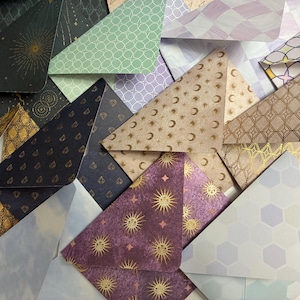 Puede incluir: Un surtido de sobres de papel plegados en varios colores y patrones. Los diseños incluyen motivos celestiales, formas geométricas y patrones florales. Los sobres son en tonos de verde, morado, azul y dorado.