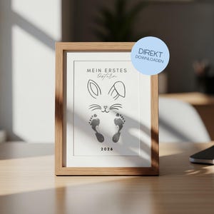 Könnte beinhalten: Ein gerahmter Druck mit hellem Holzrahmen. Der Druck zeigt ein Hasengesicht-Design mit Fußabdrücken und dem Text "MEIN ERSTES Ostern" und dem Jahr 2026. Ein blauer Kreis mit dem Text "DIREKT DOWNLOADEN" befindet sich oben rechts.