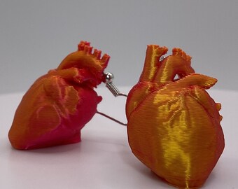 Pode incluir: Um par de brincos de coração anatómico. Os brincos são impressos em 3D com um gradiente de cores laranja e vermelho. Os brincos têm um gancho e uma mola de cor prateada.