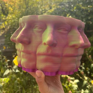 May include: A multi-faced planter in shades of pink, green, and purple. The planter features a series of human faces arranged in a circular pattern. The base of the planter is a solid purple color, and the faces have a gradient effect.