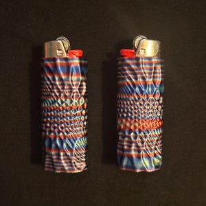 Op de afbeelding: Twee aanstekers met een textuurrijk, kleurrijk ontwerp. De aanstekers hebben een patroon van rode, blauwe en groene lijnen en stippen. De bovenkant van elke aansteker is zilverkleurig met een rode knop.