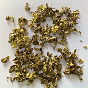 May include: A pile of small, irregularly shaped, gold-colored metal flakes on a white background.