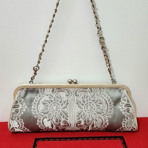 Puede incluir: Bolso de mano de satén plateado con bordado floral blanco y cadena plateada. El bolso tiene cierre de boquilla y un marco color crema. La marca "Isabella Fiore" es visible sobre una superficie roja.