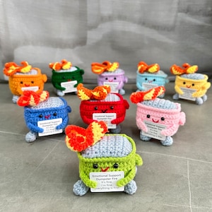 May include: A collection of ten crocheted plush toys shaped like dumpsters with flames on top. Each dumpster has a different color and a sign that reads "Emotional Support Dumpster Fire. It's fine, I'm fine. Everything is fine."