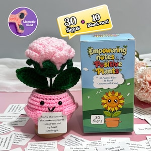 May include: A crocheted pink flower with green leaves sits in a pink pot with a smiling face. A box of "Empowering Notes by the Positive Plants" is next to it, along with cards with positive affirmations. The image also includes a magnetic hand.