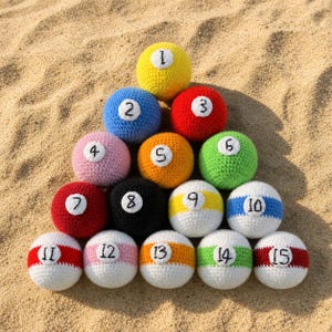 May include: A pyramid of crocheted billiard balls, each with a number from 1 to 15. The balls are in various colors, including yellow, blue, red, pink, orange, green, black, and white. The balls are arranged on a sandy surface.