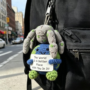 May include: A crocheted keychain featuring a gray spider atop a blue and green globe. The globe holds a sign that reads "The World Is A Better Place With You In It!" The keychain is attached to a black backpack.