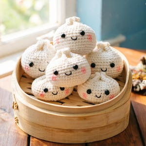 May include: A collection of handmade, crocheted dumplings with smiling faces and rosy cheeks, nestled in a bamboo steamer. Each dumpling is off-white with black eyes and a stitched smile.