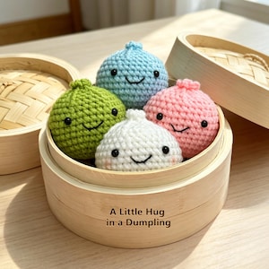 Puede incluir: Una vaporera de bambú contiene cuatro empanadillas de ganchillo en verde, azul, rosa y blanco, cada una con una cara sonriente. El texto "A Little Hug in a Dumpling" está impreso en la vaporera.