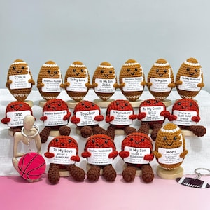 May include: A collection of handmade crochet sports-themed gifts. The assortment includes footballs and basketballs with personalized messages on small signs. The color palette features brown, orange, and white, with a pink background.