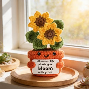 May include: Crocheted sunflower plant in an orange pot with a smiling face. The plant has yellow and brown sunflowers, green leaves, and a sign that reads "Wherever life plants you, bloom with grace."