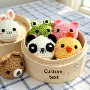 Könnte beinhalten: Ein Bambusdampfkorb mit fünf runden, gehäkelten Tierfiguren: ein grüner Frosch, ein rosa Schwein, ein weißer Panda, ein gelbes Küken und ein brauner Bär. Der Text "Custom text" ist auf der Seite des hölzernen Dampfkorbs aufgedruckt.