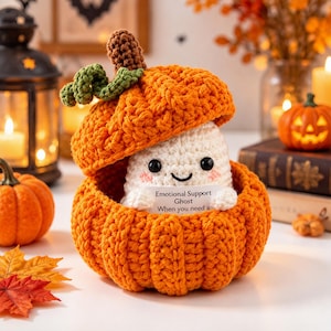 May include: A crocheted orange pumpkin with a green stem and a lid, containing a small white ghost figure with black eyes and a smile. The ghost holds a white card with the text Emotional Support Ghost When you need a hug.