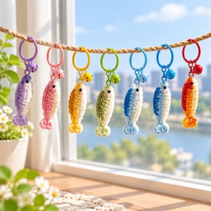 May include: A row of seven crochet fish keychains hanging from a rope. Each fish features a unique color combination, including purple, pink, yellow, green, light blue, dark blue, and orange, with a matching metal ring and a small jingle bell attached.