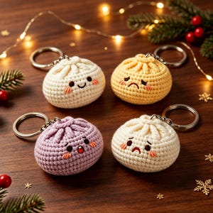 May include: Four round, crocheted dumpling keychains with embroidered facial expressions. One is white with a smile, one is yellow with a sad face, one is light purple with an open-mouthed expression, and one is cream with a frowning face.