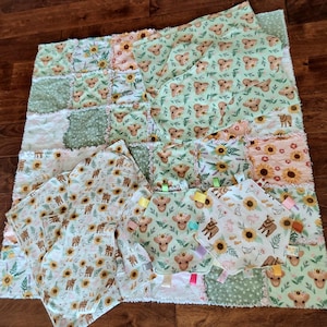 May include: A patchwork baby quilt and matching accessories. The quilt features a mix of floral and animal prints in shades of green, yellow, and peach. Several matching burp cloths and a security blanket are also visible, all with ribbon tags.