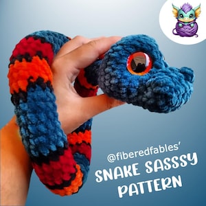May include: A crocheted snake toy with a blue and black body and red and orange stripes. The snake is curled into a circle and has a large, round eye. The text "@fiberedfables' SNAKE SASSY PATTERN" is visible in the image.