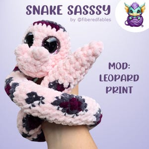 Op de afbeelding: Een roze en paarse gehaakte slangenpop met grote goggle-ogen. De slang is opgerold in een hand. De tekst "SNAKE SASSY by @fiberedfables" staat bovenaan de afbeelding. De tekst "MOD: LEOPARD PRINT" staat onderaan de afbeelding.