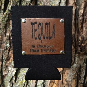 Può includere: Isolante per bevande nero con una toppa in pelle marrone. La toppa è rivettata all'isolante e presenta il testo "TEQUILA is cheaper than therapy" in lettere nere. L'isolante è progettato per contenere una lattina o una bottiglia.