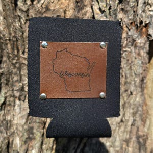 Könnte beinhalten: Schwarzer Getränkehalter mit einem braunen Lederaufnäher mit der Umrisslinie von Wisconsin und dem Wort "Wisconsin" in Schreibschrift. Der Lederaufnäher ist mit silbernen Nieten befestigt.