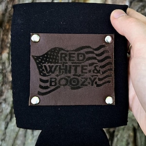 Könnte beinhalten: Ein schwarzer Getränkekühler aus Neopren mit einem rechteckigen braunen Lederaufnäher, der mit vier silbernen Metallnieten befestigt ist. Der Aufnäher ist mit einer amerikanischen Flagge und dem Text RED WHITE & BOOZY geprägt.