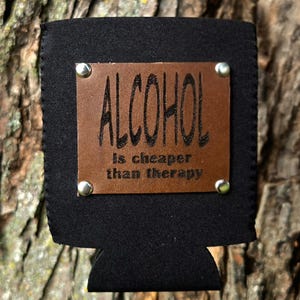 Könnte beinhalten: Schwarzer Getränkehalter mit einem braunen Lederaufnäher. Der Aufnäher ist am Halter angenietet und trägt die Worte "ALCOHOL is cheaper than therapy" in schwarzer Schrift. Der Halter ist für kalte Getränke konzipiert.