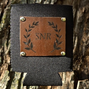 May include: Black can cooler with a brown leather patch. The patch features a laurel wreath design and the initials "SNR". The patch is secured with four silver rivets. The background is a tree trunk.