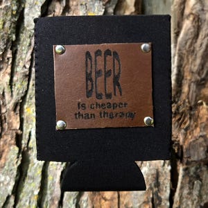 Könnte beinhalten: Schwarzer Bierdosenkühler mit einem braunen Lederaufnäher. Der Aufnäher ist am Kühler vernietet und trägt den Text "BEER is cheaper than therapy" in einer fetten, dunklen Schrift. Der Kühler ist für eine Dose in Standardgröße konzipiert.