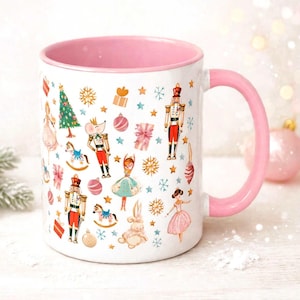 May include: A white ceramic mug with a pink interior and handle, featuring a repeating pattern of Christmas-themed illustrations including nutcrackers, ballerinas, rocking horses, gift boxes, pine trees, snowflakes, and gold stars.