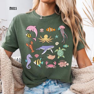 K&ouml;nnte beinhalten: Ein moosgr&uuml;nes T-Shirt mit bunten Illustrationen verschiedener Meeresbewohner, darunter ein Delfin, ein Oktopus und eine Qualle. Das Shirt hat eine lockere Passform und kurze &Auml;rmel. Das Wort "moss" ist links oben aufgedruckt.
