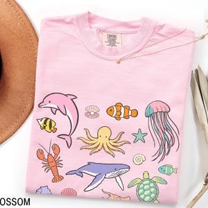 K&ouml;nnte beinhalten: Hellrosa T-Shirt mit Cartoon-Meeresbewohnern wie Delfin, Oktopus und Wal. Das Wort "BLOSSOM" ist unten links auf dem Shirt aufgedruckt.