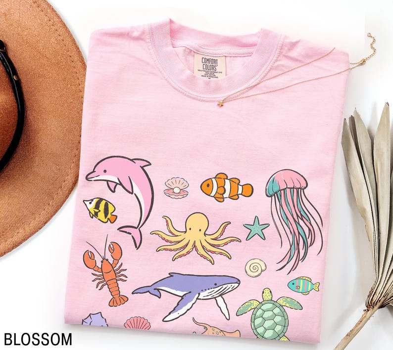 K&ouml;nnte beinhalten: Hellrosa T-Shirt mit Cartoon-Meeresbewohnern wie Delfin, Oktopus und Wal. Das Wort "BLOSSOM" ist unten links auf dem Shirt aufgedruckt.