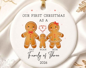 Op de afbeelding: Een ronde witte keramische kerstornament met drie peperkoekfiguren, een zuurstokhart en de tekst: OUR FIRST CHRISTMAS AS A DAD NOAH MOM Family of Three 2026. Het ornament hangt aan een goudkleurig koord.