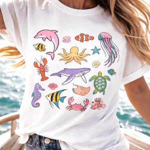 K&ouml;nnte beinhalten: Wei&szlig;es T-Shirt mit farbenfrohen Illustrationen verschiedener Meeresbewohner, darunter ein rosa Delfin, ein orangefarbener Clownfisch, ein lila Wal und eine gr&uuml;ne Schildkr&ouml;te. Weitere Meereslebewesen sind ein Seepferdchen, eine Krabbe und eine Qualle. Das Design ist skurril und verspielt.