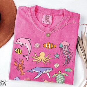 K&ouml;nnte beinhalten: Ein pinkfarbenes T-Shirt mit verschiedenen Meeresbewohnern, darunter ein Delfin, ein Oktopus und ein Wal. Das T-Shirt hat einen Rundhalsausschnitt und kurze &Auml;rmel. Eine goldene Halskette mit Anh&auml;nger liegt auf dem T-Shirt. Der Text "Crunch Berry" steht unten links.