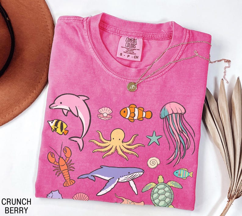 K&ouml;nnte beinhalten: Ein pinkfarbenes T-Shirt mit verschiedenen Meeresbewohnern, darunter ein Delfin, ein Oktopus und ein Wal. Das T-Shirt hat einen Rundhalsausschnitt und kurze &Auml;rmel. Eine goldene Halskette mit Anh&auml;nger liegt auf dem T-Shirt. Der Text "Crunch Berry" steht unten links.