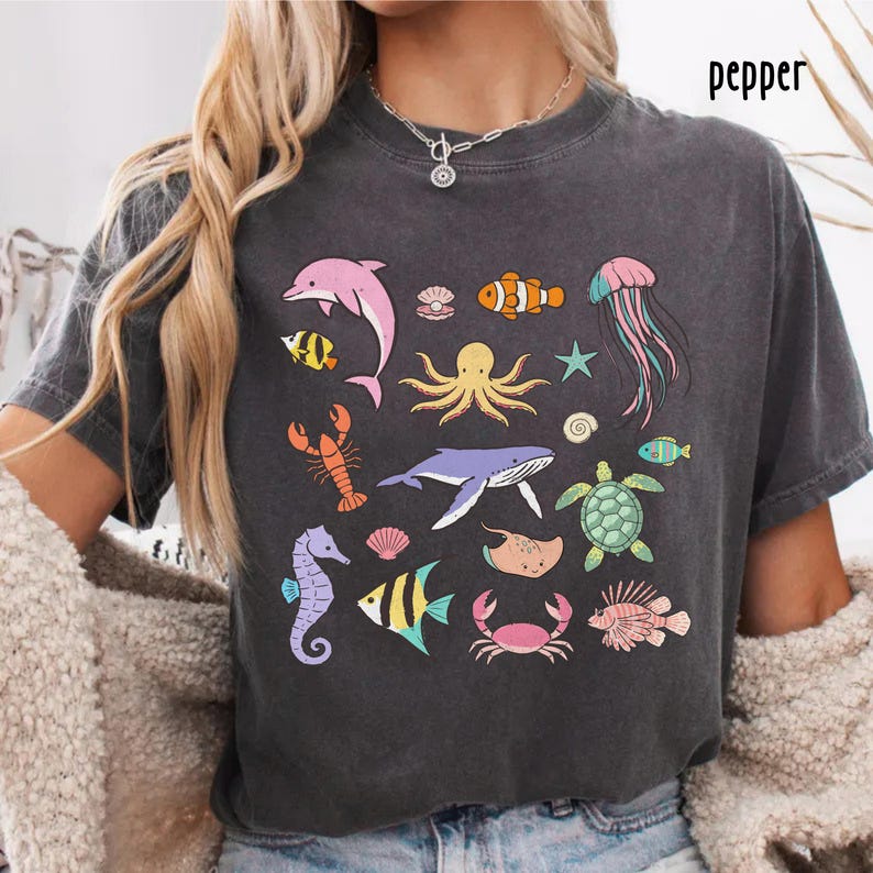 K&ouml;nnte beinhalten: Dunkelgraues T-Shirt mit farbenfrohen Illustrationen verschiedener Meeresbewohner, darunter ein rosa Delfin, ein orangefarbener Clownfisch und ein lila Oktopus. Das Wort "pepper" ist oben rechts aufgedruckt. Das Design umfasst ein Seepferdchen, eine Krabbe und eine Qualle.