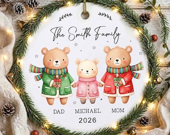 Op de afbeelding: Een rond wit keramieken ornament met een illustratie van drie beren met sjaals en jassen die cadeaus vasthouden. De tekst luidt The Smith Family, Dad, Michael, Mom en 2026. Het ornament is omlijst door een groene dennenkrans.