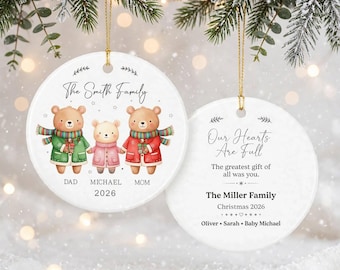 May include: Two round white ceramic ornaments with gold hanging strings. One features three illustrated bears in winter clothing with the text The Smith Family, Dad, Michael, 2026. The other features the text Our Hearts Are Full, The Miller Family, Christmas 2026, and Oliver, Sarah, Baby Michael.