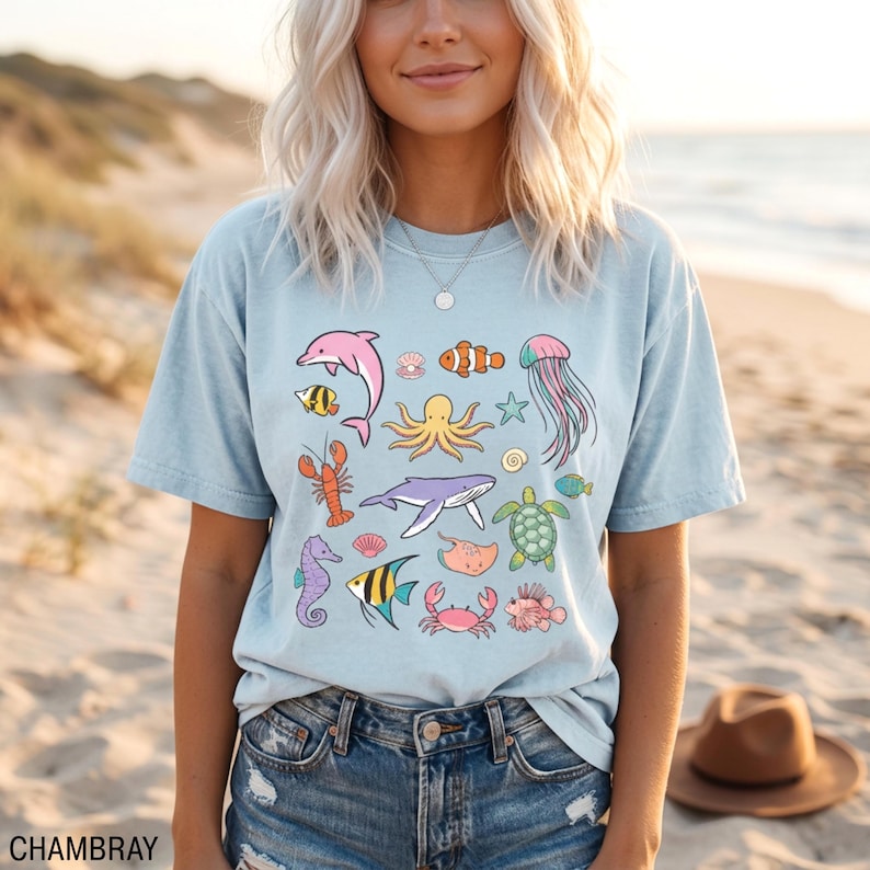 K&ouml;nnte beinhalten: Hellblaues T-Shirt mit einer farbenfrohen Meereslebewesen-Grafik. Das Design umfasst einen rosa Delfin, einen Oktopus, einen Wal und verschiedene andere Meeresbewohner. Das Wort "CHAMBRAY" ist unten links aufgedruckt.