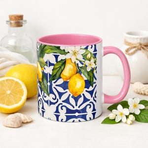 May include: A white ceramic mug with a pink interior and handle. The mug features a blue and white tile pattern with a lemon and floral design. A sliced lemon and whole lemon are next to the mug.