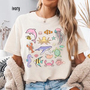 K&ouml;nnte beinhalten: Elfenbeinfarbenes T-Shirt mit bunten Cartoon-Meeresbewohnern, darunter ein rosa Delfin, ein Oktopus, eine Qualle und verschiedene Fische. Das Wort "ivory" ist oben links auf dem Shirt aufgedruckt. Das Shirt ist ein l&auml;ssiger, bequemer Stil.