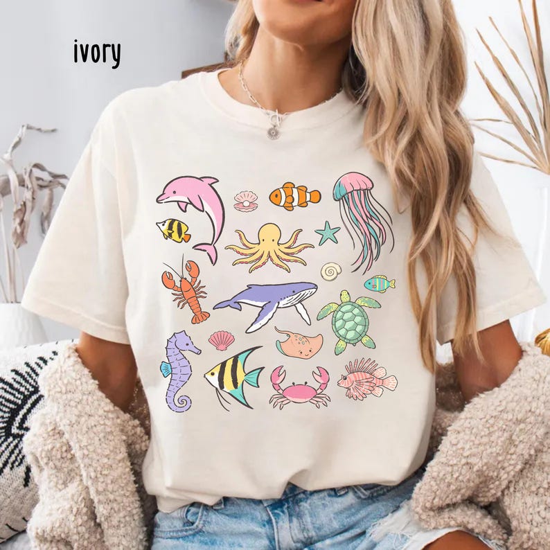 K&ouml;nnte beinhalten: Elfenbeinfarbenes T-Shirt mit bunten Cartoon-Meeresbewohnern, darunter ein rosa Delfin, ein Oktopus, eine Qualle und verschiedene Fische. Das Wort "ivory" ist oben links auf dem Shirt aufgedruckt. Das Shirt ist ein l&auml;ssiger, bequemer Stil.