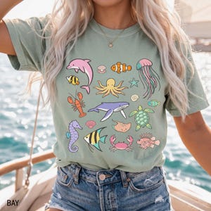 K&ouml;nnte beinhalten: Salbeigr&uuml;nes T-Shirt mit bunten Cartoon-Meeresbewohnern, darunter ein Delfin, ein Oktopus und eine Qualle. Das Shirt hat eine lockere Passform und einen Rundhalsausschnitt. Das Wort "BAY" ist unten links aufgedruckt.