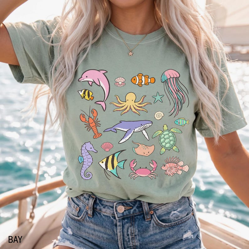 K&ouml;nnte beinhalten: Salbeigr&uuml;nes T-Shirt mit bunten Cartoon-Meeresbewohnern, darunter ein Delfin, ein Oktopus und eine Qualle. Das Shirt hat eine lockere Passform und einen Rundhalsausschnitt. Das Wort "BAY" ist unten links aufgedruckt.