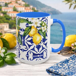 May include: White ceramic mug with a blue interior and handle. The mug features a lemon and floral design with a blue and white pattern. Yellow lemons and sunglasses are next to the mug.