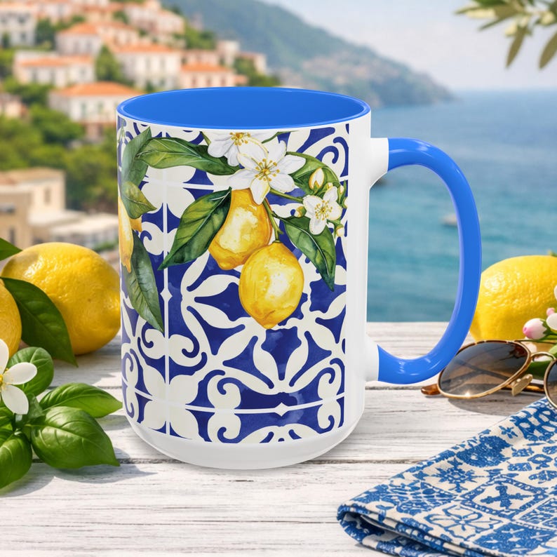 May include: White ceramic mug with a blue interior and handle. The mug features a lemon and floral design with a blue and white pattern. Yellow lemons and sunglasses are next to the mug.