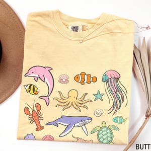 K&ouml;nnte beinhalten: Hellgelbes T-Shirt mit bunten Meeresbewohnern, darunter ein rosa Delfin, ein orangefarbener Clownfisch und ein blauer Wal. Das Shirt hat einen Rundhalsausschnitt und kurze &Auml;rmel. Der Text "COMFORT COLORS" ist sichtbar.