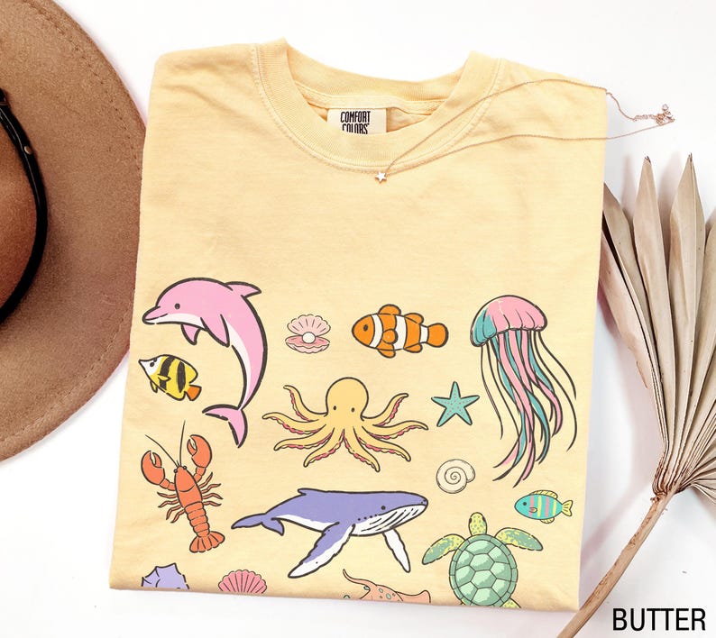 K&ouml;nnte beinhalten: Hellgelbes T-Shirt mit bunten Meeresbewohnern, darunter ein rosa Delfin, ein orangefarbener Clownfisch und ein blauer Wal. Das Shirt hat einen Rundhalsausschnitt und kurze &Auml;rmel. Der Text "COMFORT COLORS" ist sichtbar.